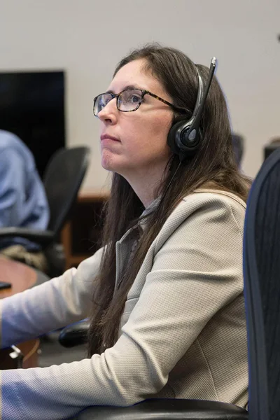 A woman with long dark hair, glasses, and a headset looks intently upward. She wears a beige blazer.